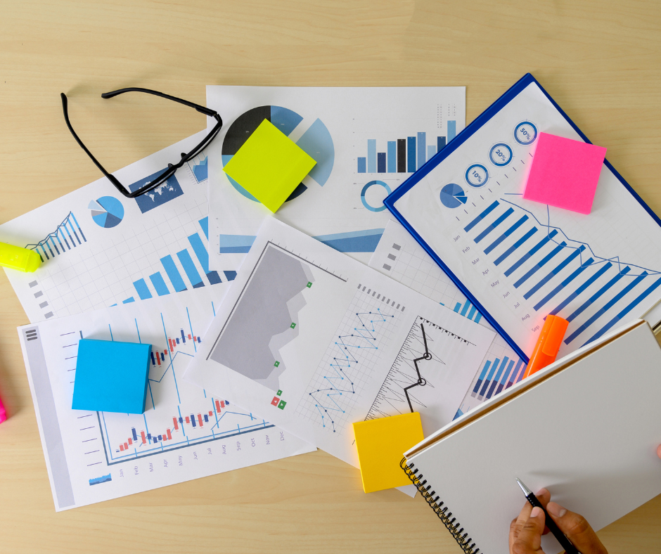 5 messily spread out sheets of paper with blue graphs, post it notes on top of the graphs, glasses on top of paper, and a hand taking notes on a blank notebook.