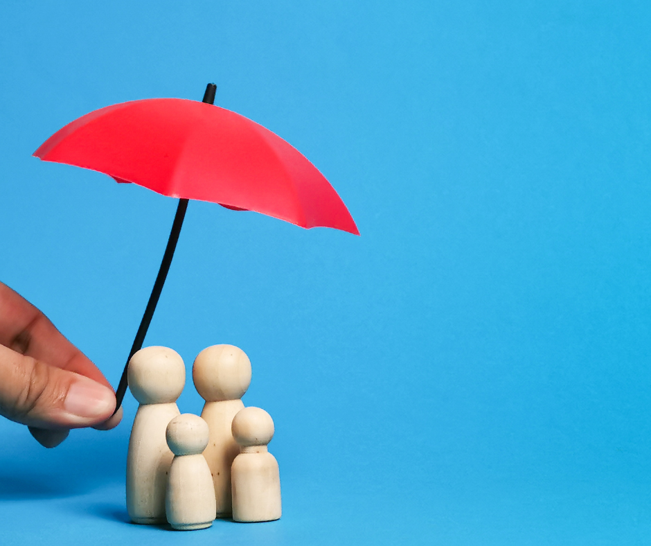 A family of 4 wooden people covered by a mini red umbrella held by a human.