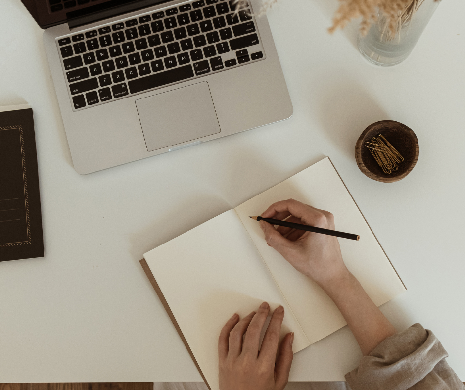 Top-down view of person writing in an empty notebook with a black pen and a laptop in front of them.