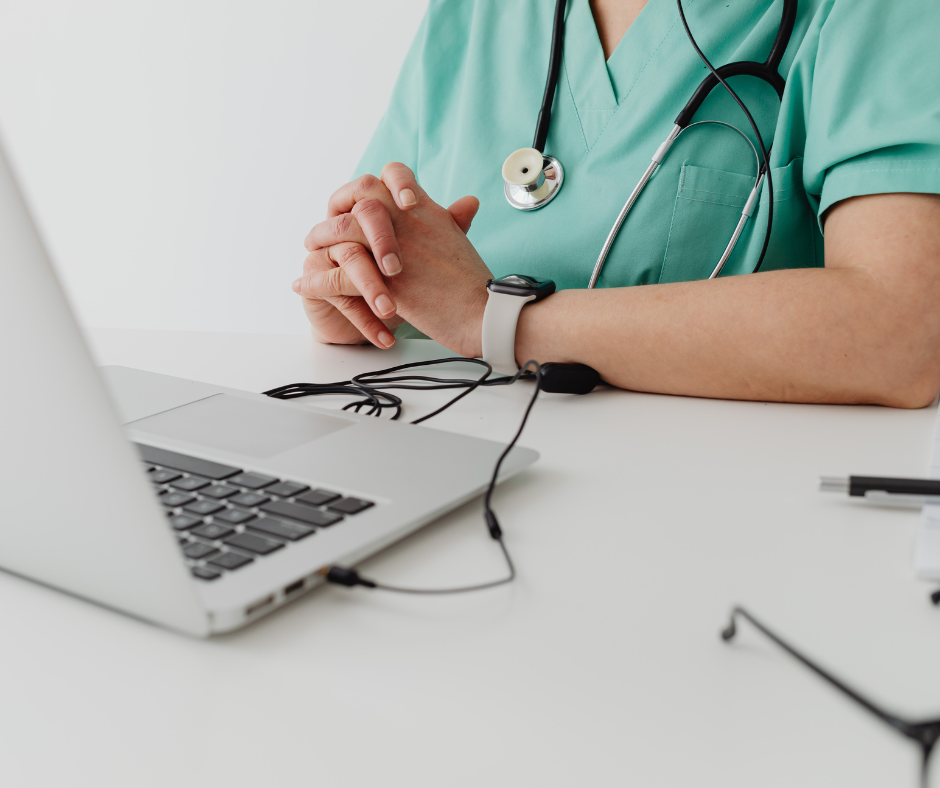 Person in teal scrubs and wearing a stethoscope around neck with earbuds facing silver laptop.