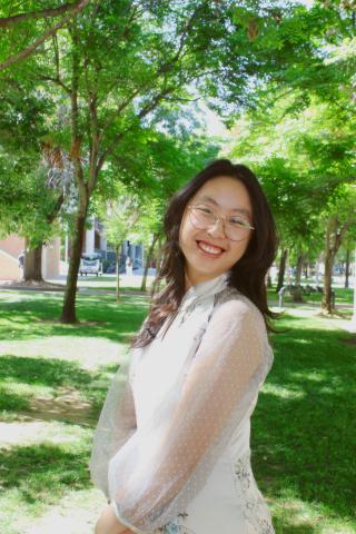 Woman with black hair wearing a white dress in front of green trees