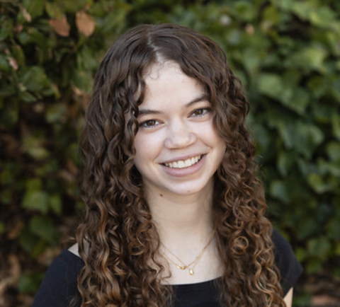 Woman with brown curly hair in front of foliage. 