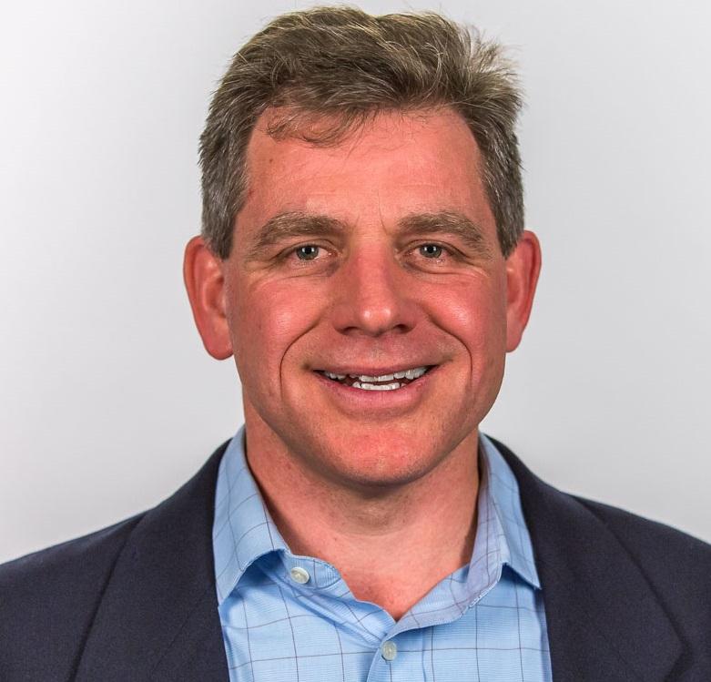 Caucasian man in a blue patterned button down wearing a black suit jacket with brown-gray hair and dark gray eye color.