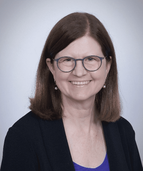 Smiling Caucasian adult woman with straight shoulder length brown hair, cobalt eyes, black glasses, and earrings wearing a dark blue blouse and black blazer.