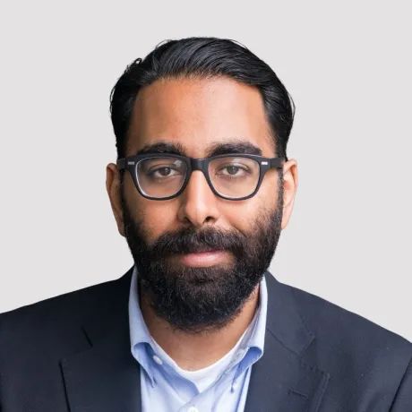 Indian man with black hair slick back hair and a beard wearing a blue button down and black suit jacket.